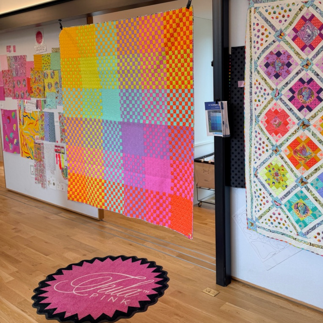 Colorful quilts on display in a room with a pink and black logo on the floor.